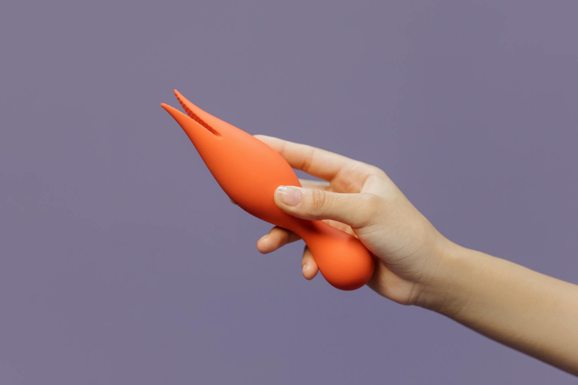Hand hält einen modernen orangefarbenen Vibrator gegen minimalistischen violetten Hintergrund