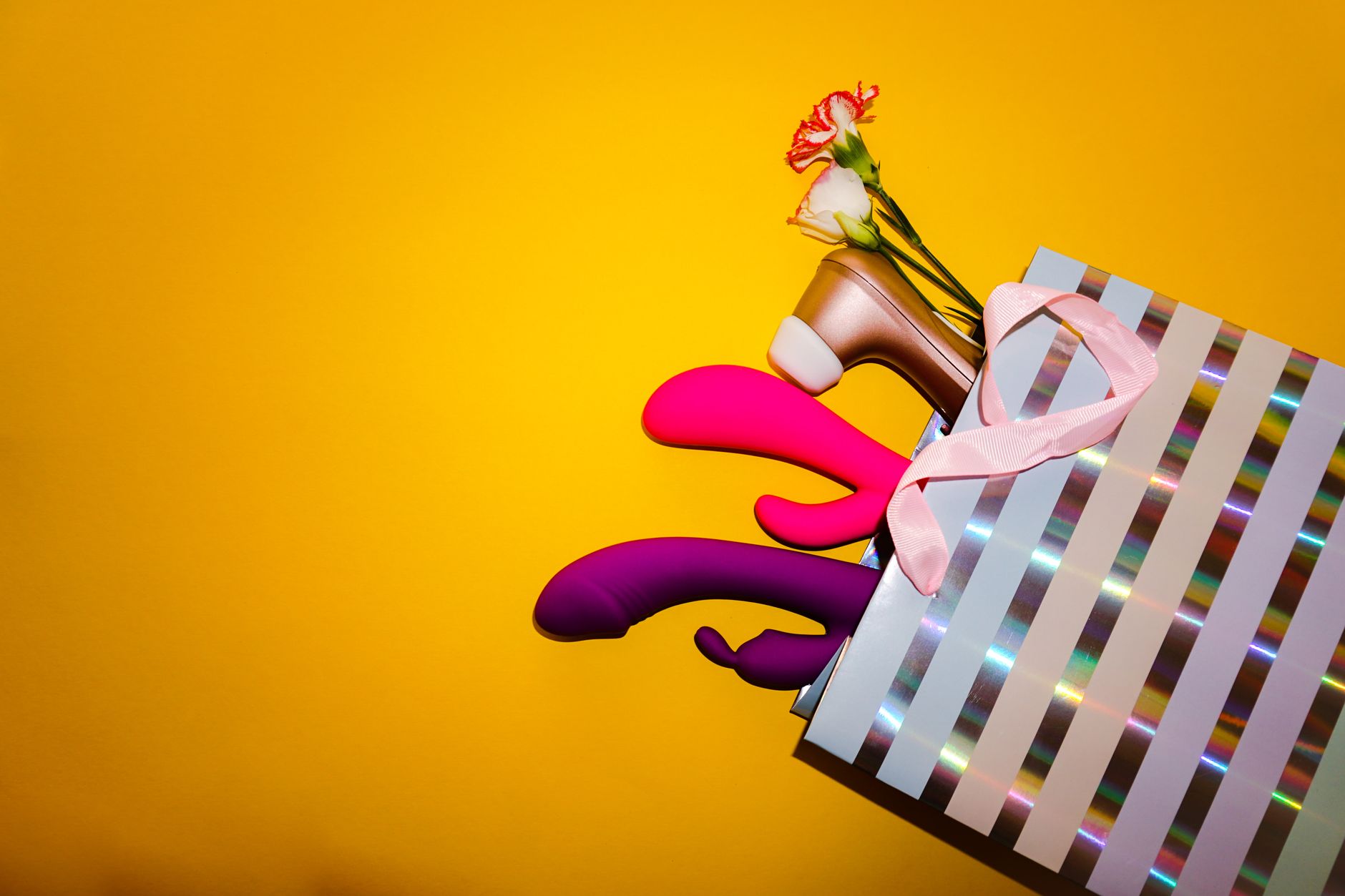 Colorful vibrators with flowers in a holographic gift bag against a bold yellow background